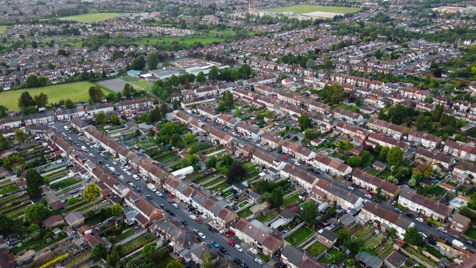 An aerial view of a residential suburban area with rows of terraced houses featuring small front gardens, some with lush green lawns, flower beds, and paved pathways. The streets are lined with parked